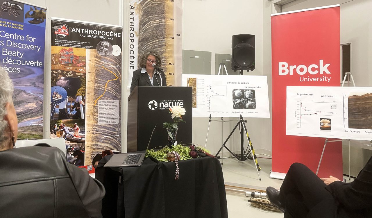 Brock University Acting Vice-President, Research Michelle McGinn looks down as she stands at a podium with the name and logo of the Canadian Museum of Nature. In the background are a variety of pop-up banners, including one from Brock University, and charts depicting scientific graphs. 