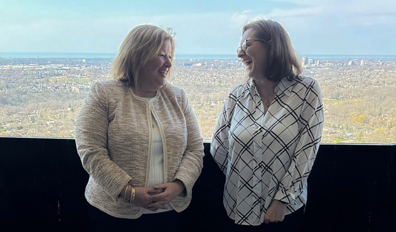 Ontario’s Minister of Agriculture, Food and Rural Affairs Lisa Thompson and Brock President and Vice-Chancellor Lesley Rigg smiling on a balcony on a sunny day, the Niagara region at a distance in the background.
