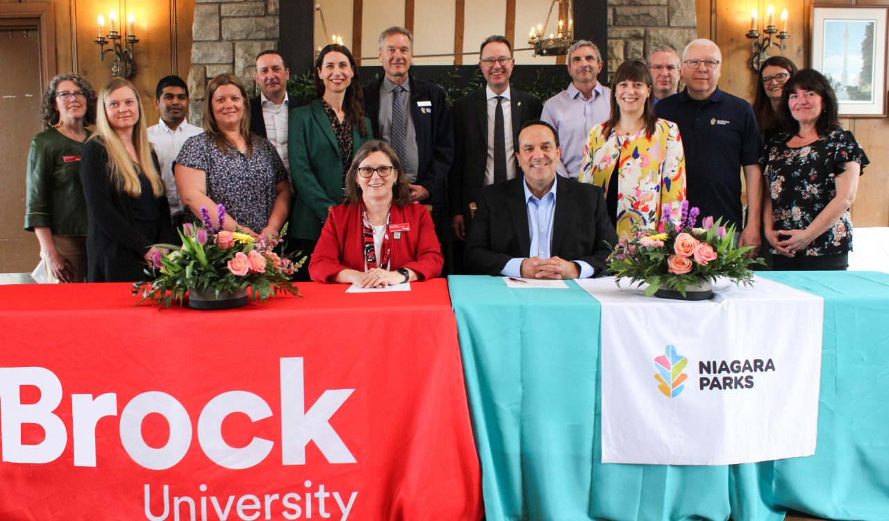 A large group of people stand behind two people seated at tables with Brock University and Niagara Parks table cloths. 