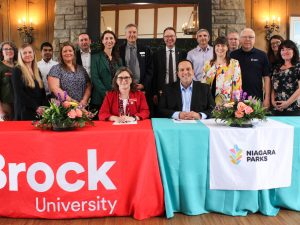A large group of people stand behind two people seated at tables with Brock University and Niagara Parks table cloths.