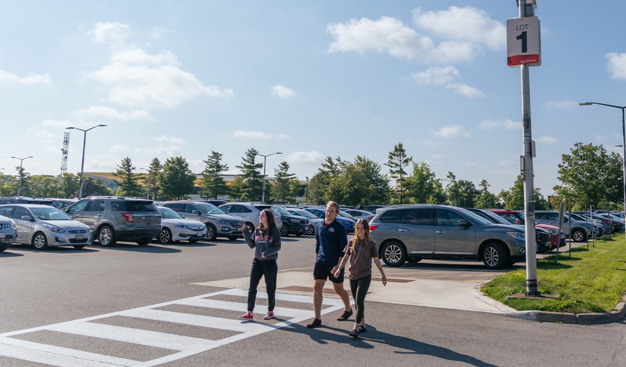 Three students walk towards campus from the Lot 1 parking lot with cars in the background.