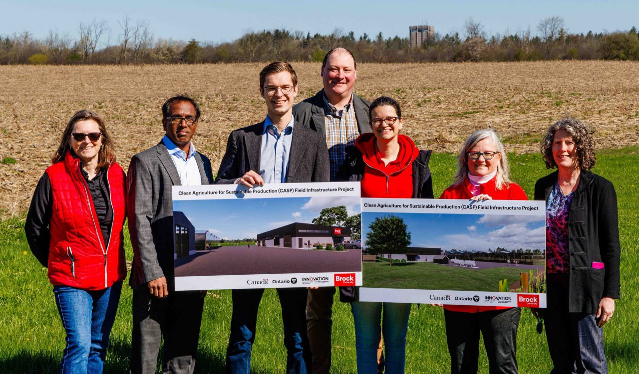 Seven people stand in a farm field, two of them holding renderings of a research farm to be built on the property. 