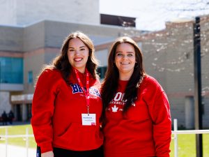 Two women pose in red sweaters outside in the sun at Brock University’s main campus.