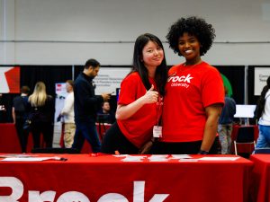 Two women in red T-shirts pose inside of a gymnasium at Brock University.