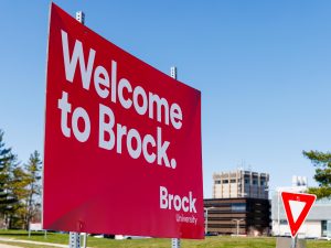 A bright red sign shines under a blue sky outside of Brock University.