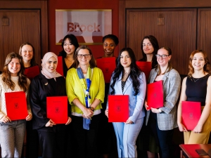 A group of 11 women stand smiling in a boardroom with awards in red folders from Brock University.