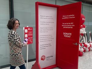A woman taps her cellphone on a scanner beside a red door displayed in a bright atrium.