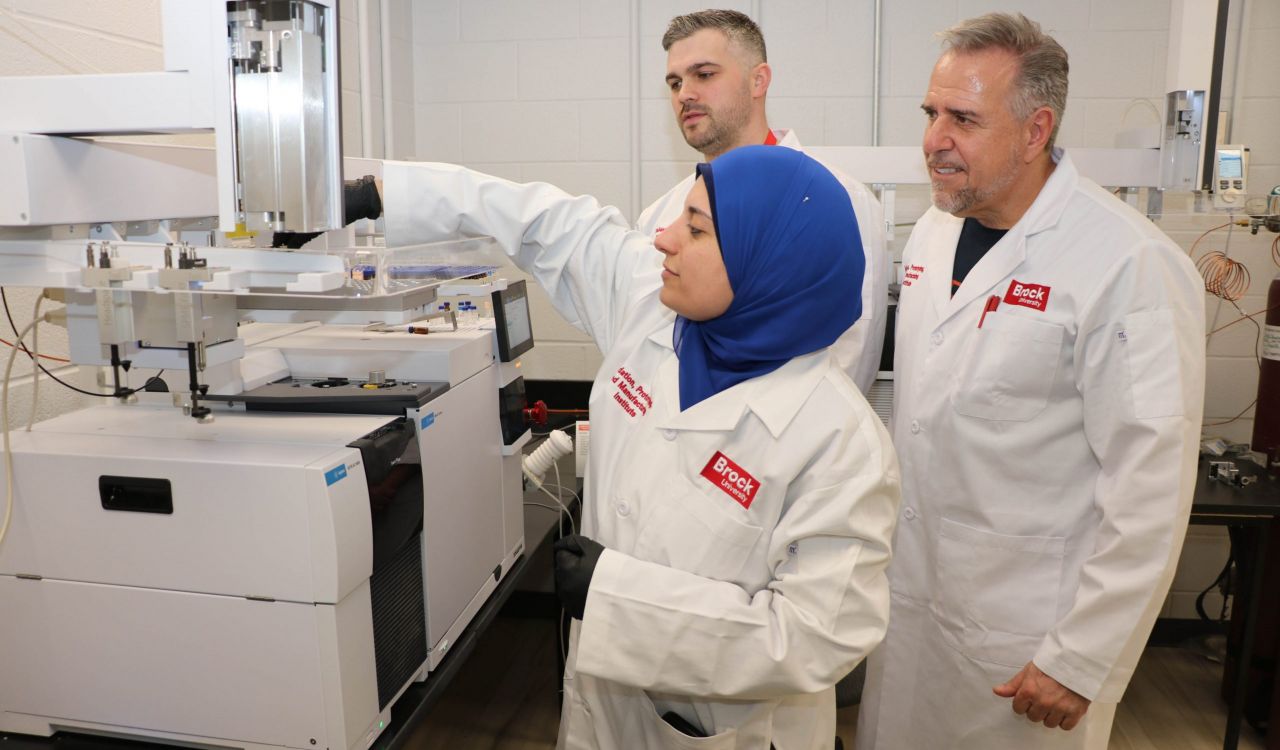 Research Associate Noha Morsy (centre) wearing a blue headscarf stretches out her arm over a white machine to place a vial in a section on top of the machine. Standing right behind her are VPMI Instrument Technologist Dave Bowman and VPMI Business Director Ivano Labricciosa