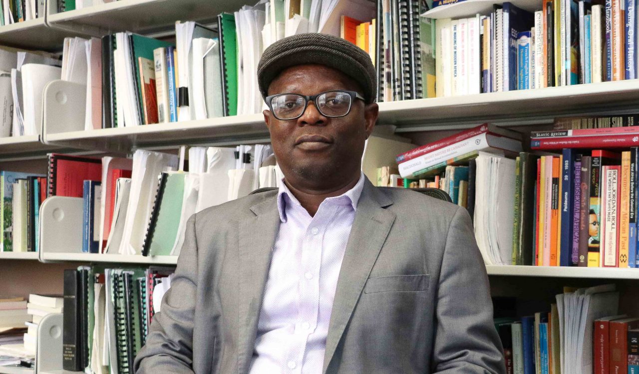 Brock History Professor Olatunji Ojo sits in front of bookcase filled with colourful books. He wears glasses and a hat and grey blazer, hands folded in his lap will intently looking at the camera.