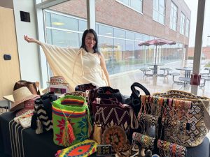 Colombian Wayüü Indigenous artist Yeimis Monsalve stands at Brock University in front of a window showing a rainy day. She wears a cream-coloured dress with her hand outstretched, showing a table of colourful bags, hats and bracelets on display.