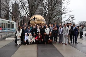 A diverse group of 37 university students pose in front of a bronze globe statue outside the United Nations building in New York City.