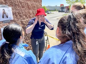 A woman in a red hat holds her hands over her ears while speaking with nursing students in blue scrubs during an emergency event simulation in a parking lot.