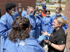 A group of nursing students in blue gowns stand around an instructor during an emergency event simulation in a parking lot.