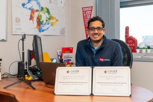 A man sits behind a desk in an office posing for a photo with two certificates.