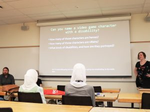 A teacher stands at the front of a classroom beside a projector screen, with students seated at tables looking on.