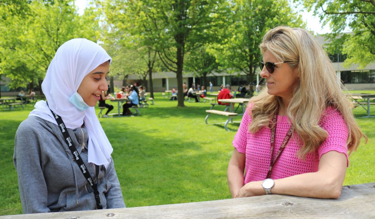 A blind female student sits with a teacher at a picnic table on a university campus.