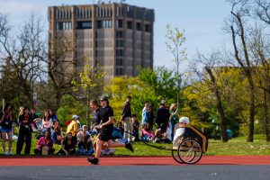 Two girls pull a boy in a chariot on a track while fellow students cheer them on a sunny afternoon at Canada Games Park.