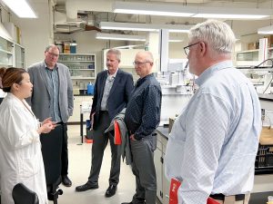 A woman in a lab coat speaks with a group of men in a lab.