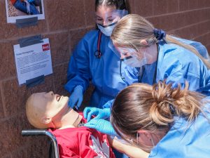 Three nursing students in blue gowns assess a mannequin patient in a wheelchair.
