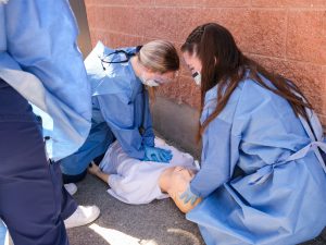 Two nursing students in blue gowns practice CPR on a mannequin patient.
