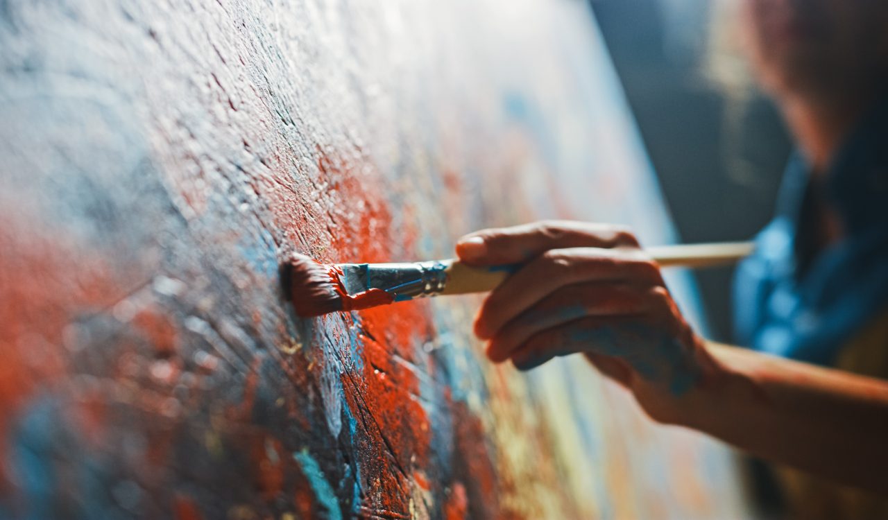 A close-up of a hand holding a paintbrush against a colourful canvas.