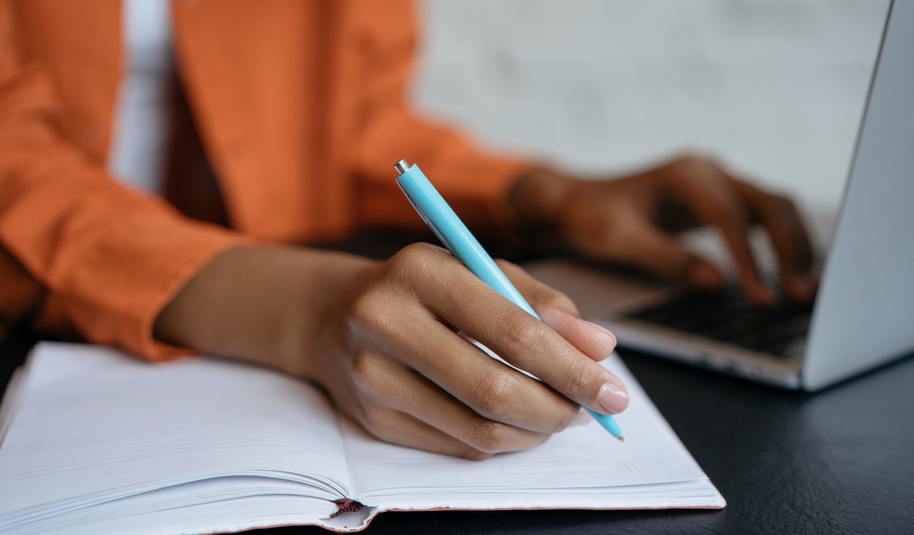 Close-up of hands, with on gripping a pen writing in a notebook and in the background fuzzed out the other hand typing on a laptop keyboard.