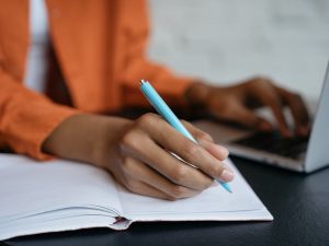 Close-up of hands, with on gripping a pen writing in a notebook and in the background fuzzed out the other hand typing on a laptop keyboard.