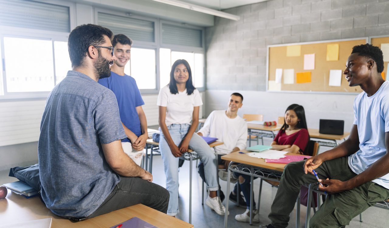 A diverse group of teenage students sitting on desks in a high school classroom; in the foreground are two young men looking at each other, smiling, with two young men and women smiling as they watch the two men talking.