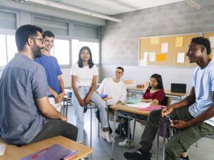 A diverse group of teenage students sitting on desks in a high school classroom; in the foreground are two young men looking at each other, smiling, with two young men and women smiling as they watch the two men talking.