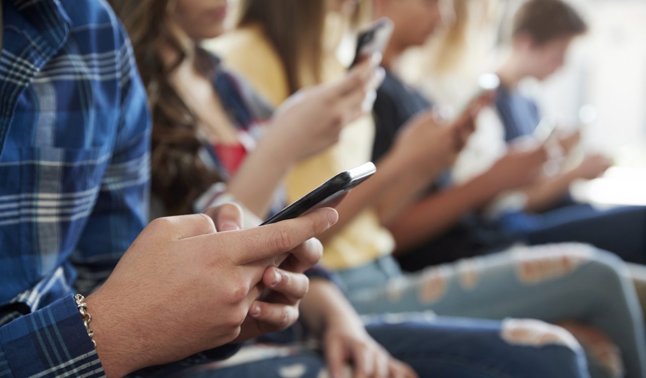 A close-up of students sitting in a line, each looking at a cellphone in their hands.