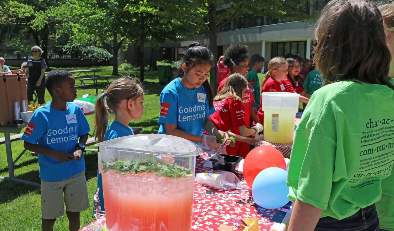 Students stand around a table selling lemonade to a customer.