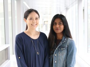 Mid-range view of third-year biological sciences student Paige Au (left) and fourth-year biochemistry student Raahavi Ramathesun (right) standing side-by-side with a fuzzy hallway in the background.