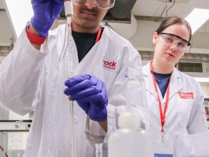 A high school student wearing a laboratory coat, safety glasses and latex gloves dips a long glass pipette into a beaker of cola as a fellow student looks over their shoulder.