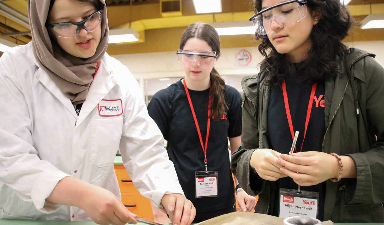 A young woman dressed in a white laboratory coat wipes off a scientific instrument with a paper towel. To the right, two high school girls wearing name tags for Brock University’s Scientifically Yours conference look down at the instrument.