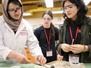 A young woman dressed in a white laboratory coat wipes off a scientific instrument with a paper towel. To the right, two high school girls wearing name tags for Brock University’s Scientifically Yours conference look down at the instrument.