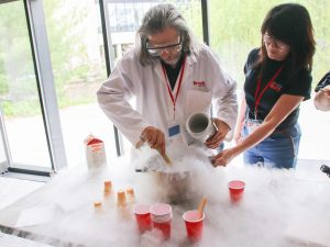 An older man wearing a laboratory coat and safety glasses stirs a large pot of ice cream. Fog encircles the pot and is escaping from a container of liquid nitrogen he holds in his other hand. A woman to the right assists by holding the handle of the pot.