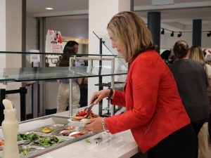 A woman in a red blazer, uses tongs to put red peppers on her plate from a salad bar.