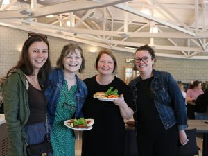 Four standing women smile and pose for the camera in a campus cafeteria.