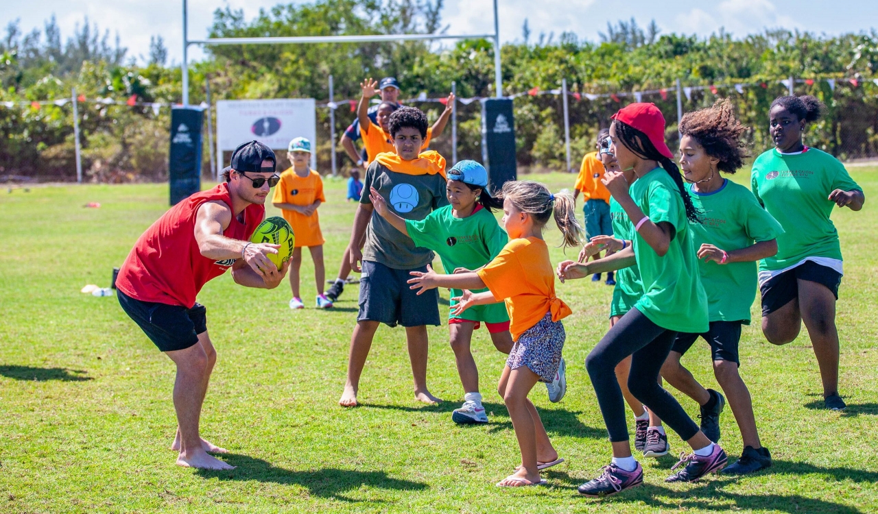 A young adult coach holds a rugby ball as several children hold out their hands and approach it.