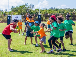 A young adult coach holds a rugby ball as several children hold out their hands and approach it.