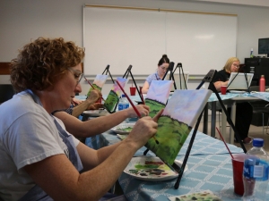 A woman in a classroom sits at a table and paints a canvas placed on an aisle. In the background two more women can be seen doing the same.