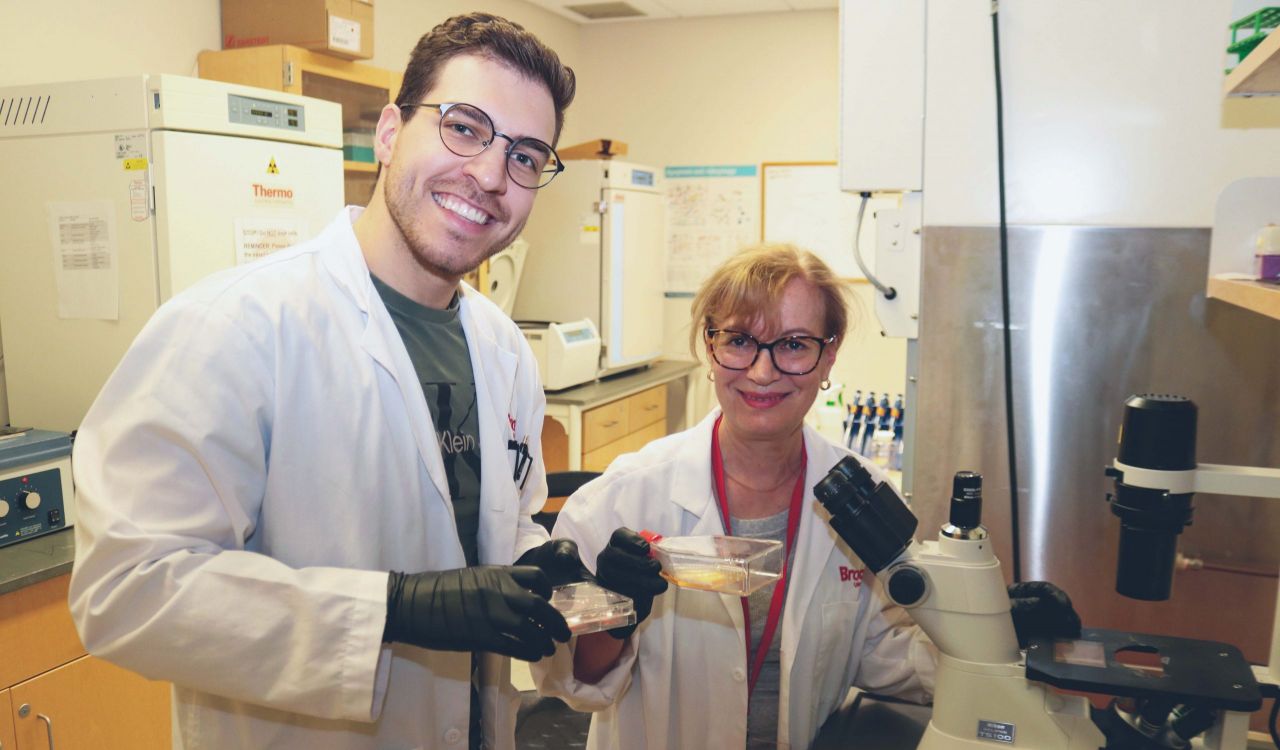 Close up of master’s student Matteo Nadile (left) and Professor of Health Sciences Evangelia Tsiani (right) smiles into the camera as they stand next to a microscope, with other lab machines and benches in the background. Each person is holding a dish filled with liquid and wearing black gloves.