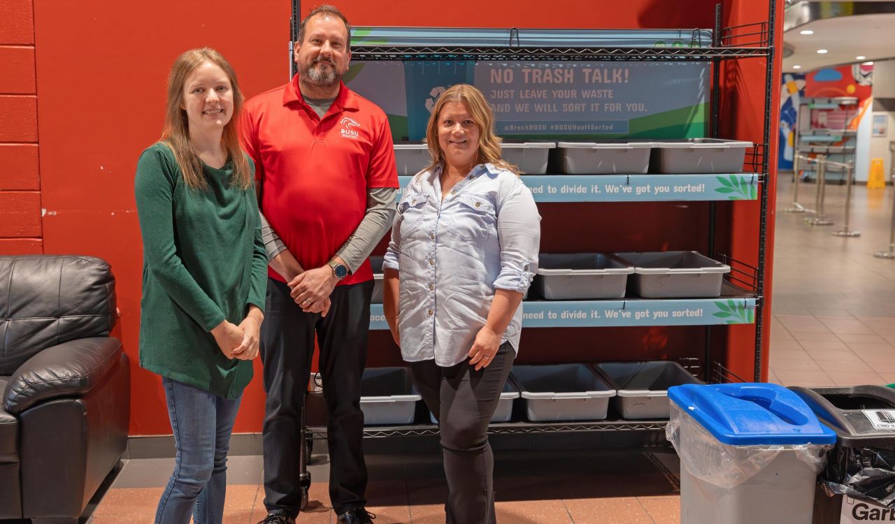 Three people stand together in front of a series of plastic bins on a rack.