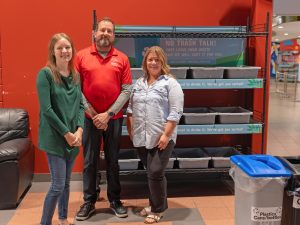 Three people stand together in front of a series of plastic bins on a rack.