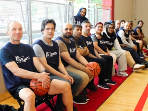 A group of people wearing basketball jerseys sit courtside at a basketball game on a team bench.