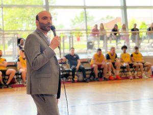 A man speaks with a microphone in front of a group of basketball players in a gymnasium.