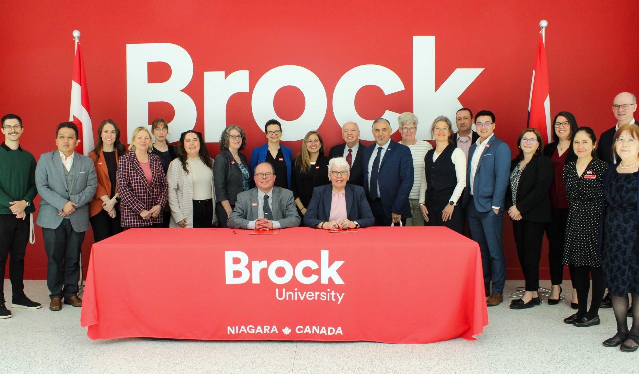 A large group of people pose in front of a red wall with the word Brock on it in white. Two of the people are sitting at a table with a red Brock University cloth on it.