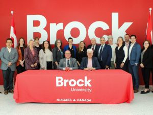 A large group of people pose in front of a red wall with the word Brock on it in white. Two of the people are sitting at a table with a red Brock University cloth on it.