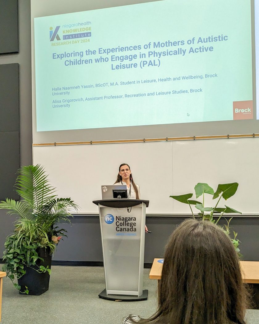 A woman stands at a podium that reads “Niagara College” with a large screen behind her projecting a presentation on “Exploring the Experiences of Mothers of Autistics Children who Engage in Physical Active Leisure (PAL).