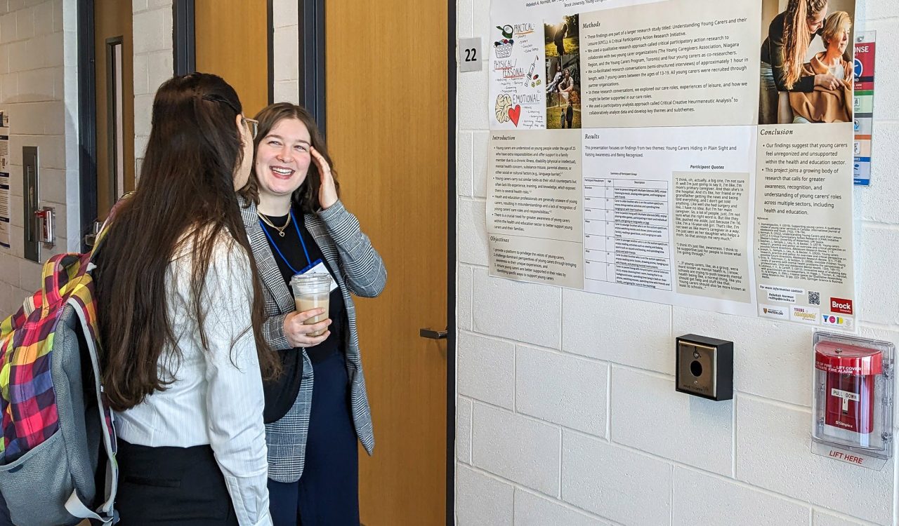 Two women stand by a research poster taped to a white wall. One woman is smiling; she touches her face with her left hand and holds a cup of coffee with her right hand. The face of the woman she is speaking with is not seen; only the back of her head is visible.